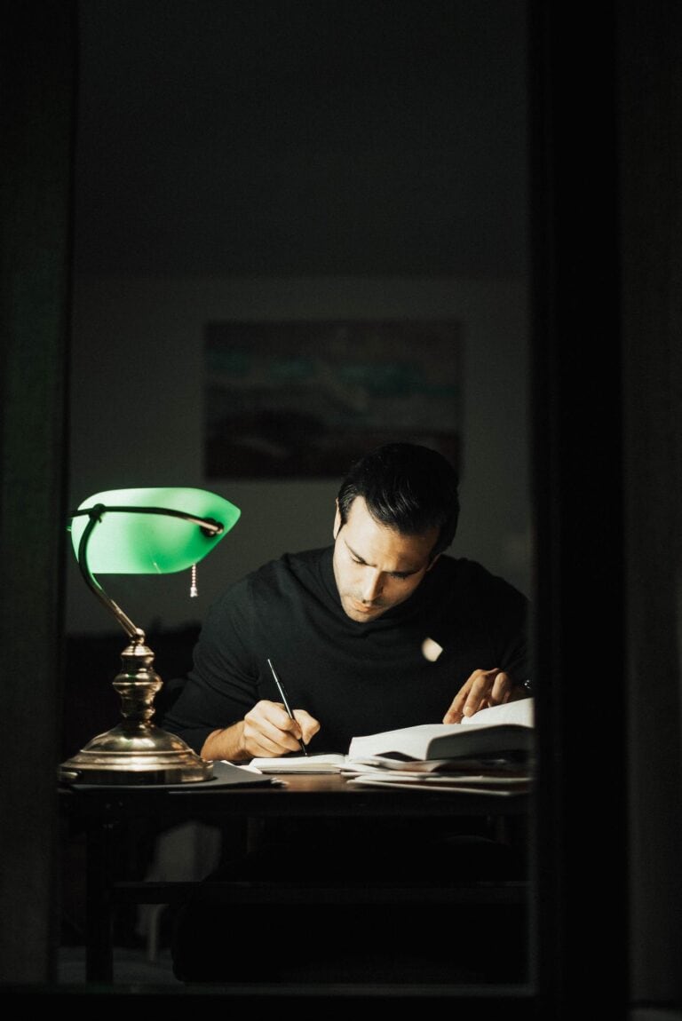 Writing a Novel ~ Concentrated young male wearing black turtleneck taking notes and reading books while sitting at desk with papers and bankers lamp in dark home office