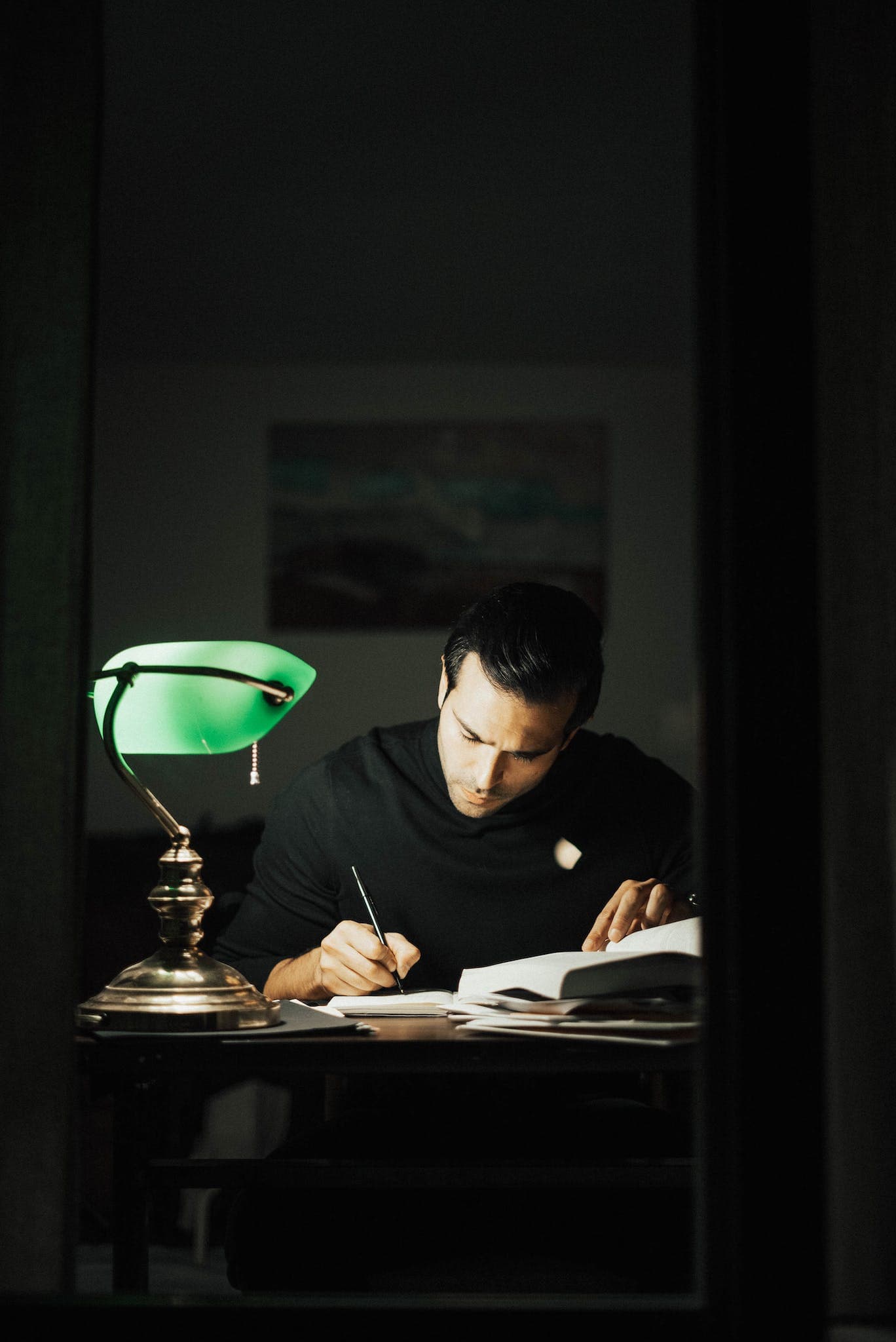 Writing a Novel ~ Concentrated young male wearing black turtleneck taking notes and reading books while sitting at desk with papers and bankers lamp in dark home office