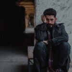 Despaired ethnic man in dark clothes touching head and sitting on stone window opening in shabby dusty building