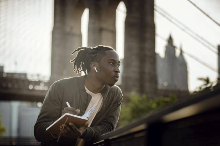 Writer's Block ~ A Positive black man listening to music with wireless earphones and taking notes in diary on street but has a slight Writer's Block