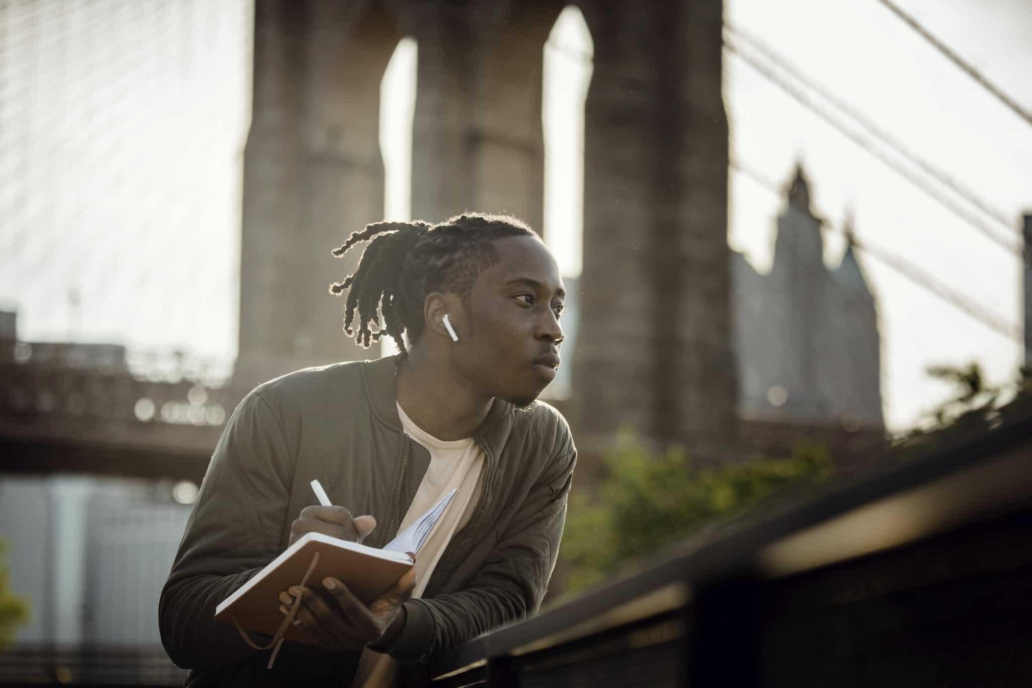 Writer's Block ~ A Positive black man listening to music with wireless earphones and taking notes in diary on street but has a slight Writer's Block
